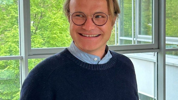 Mann in blauer Strickjacke und Jeans lächelt in einem Raum mit großen Fenstern.