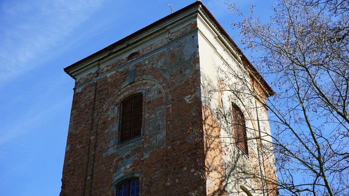 Backsteinturm mit Fensteröffnungen und geschlossener Holzjalousie vor blauem Himmel und kahlem Baum