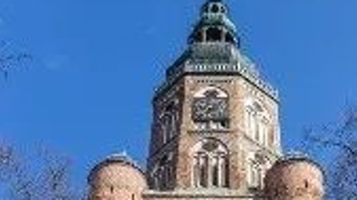 Historischer Backsteinturm mit Uhr und dekorativer Spitze vor klarem blauem Himmel.