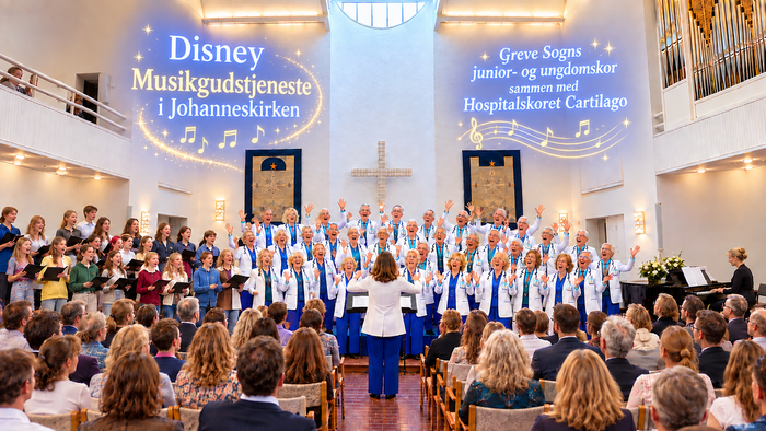 Et stort kor optræder i en kirke med publikum og bannere, der fejrer Disney Musikgudstjeneste og arrangementet Greve Sogn.
