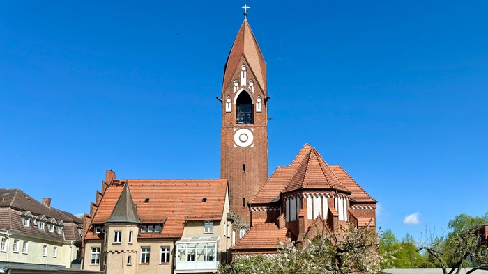 Bild zeigt eine Kirche mit hohem Turm und Uhr, umgeben von einem gepflegten Garten.