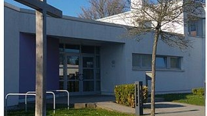 Das Bild zeigt ein modernes Gebäude mit einem blauen Eingangstor und einem kleinen Baum davor.