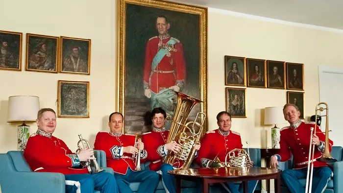 Uniformerede musikere poser med messinginstrumenter i et rum prydet med historiske portrætter og et fremtrædende portræt over dem.