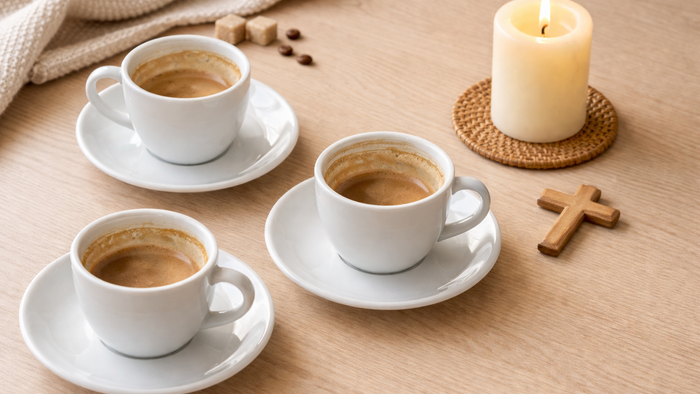 Hyggelig opsætning med tre kopper cappuccino, et stearinlys, kaffebønner og et trækors på et rustikt bord.