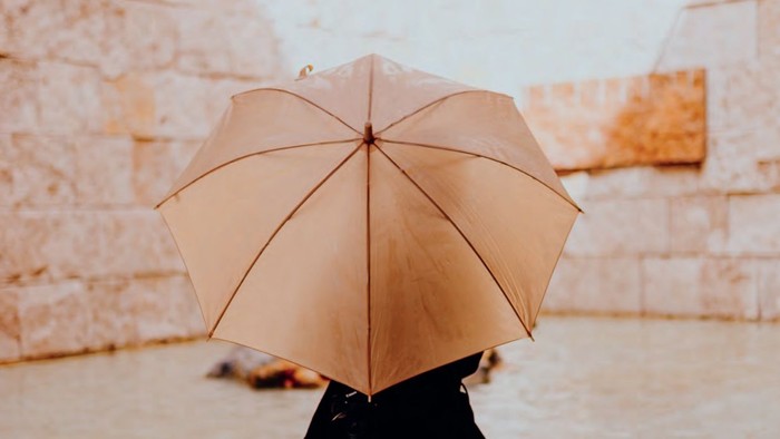 Eine Person hält einen hellbraunen Regenschirm vor einem steinernen Mauerhintergrund, möglicherweise an einem historischen oder antiken Ort.