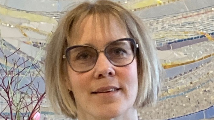 En person med kort, blondt hår og briller iført en mønstret bluse foran en farverig mosaikvæg.