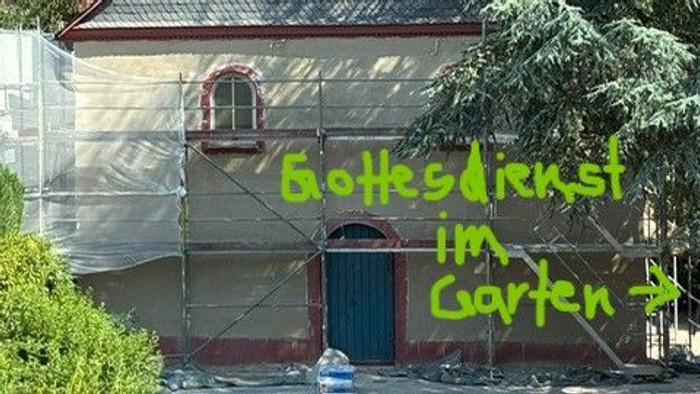 Eine Kirche im Umbau mit Gerüsten, die mit dem Schild „Gottesdienst im Garten“ auf Freiluftgottesdienste hinweist.