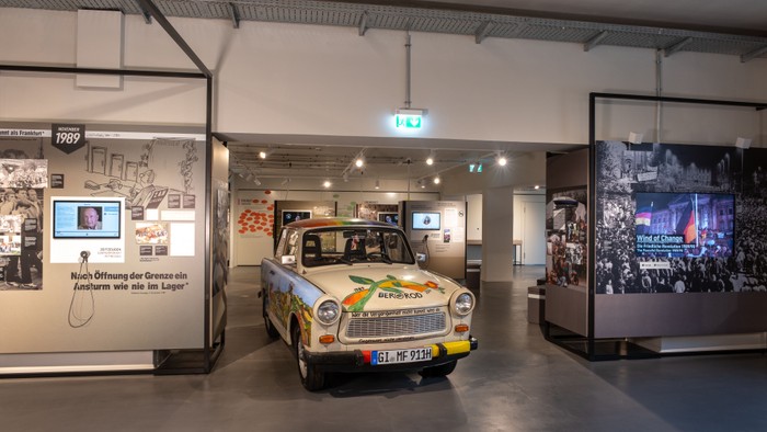 Eine Museumsausstellung mit einem farbenfrohen, historischen Trabant-Auto, umgeben von informativen historischen Schautafeln.