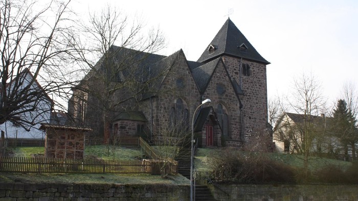 Steinerne Kirche mit spitzem Dach und kleinem Turm, umgeben von kahlen Bäumen und einer niedrigen Steinmauer.