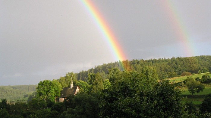 Eine friedvolle Landschaft mit einem doppelten Regenbogen, der sich über üppige grüne Hügel und ein idyllisches Haus wölbt.
