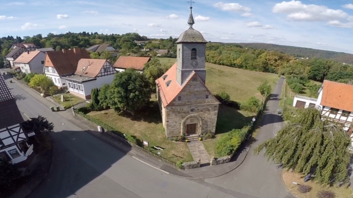 Luftaufnahme eines malerischen Dorfes mit einer kleinen Steinkirche, umgeben von Häusern und grünen Feldern.