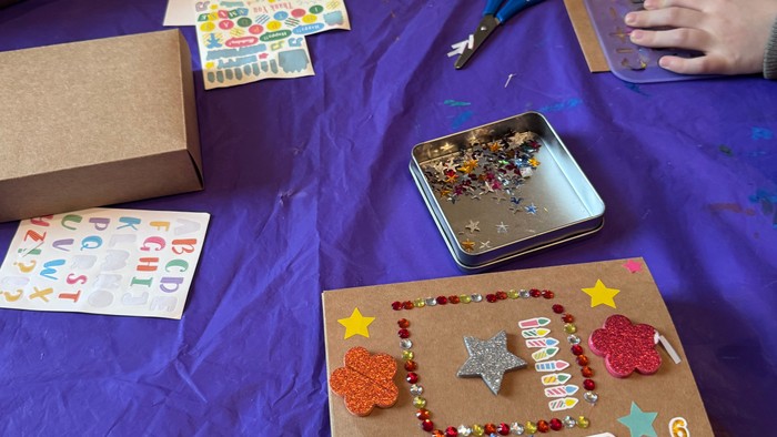 Kinder, die eine festliche Geburtstagskiste mit Aufklebern, Glitzer und Dekorationselementen auf einem lila Tischtuch basteln.