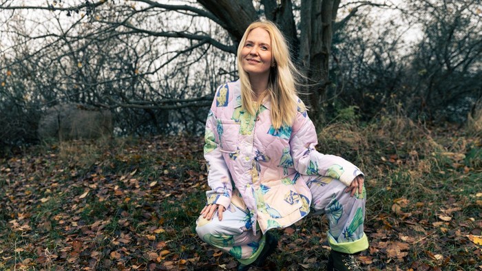En glad kvinde i blomstrede pyjamas sidder lykkeligt på en skovbund omgivet af faldne blade.