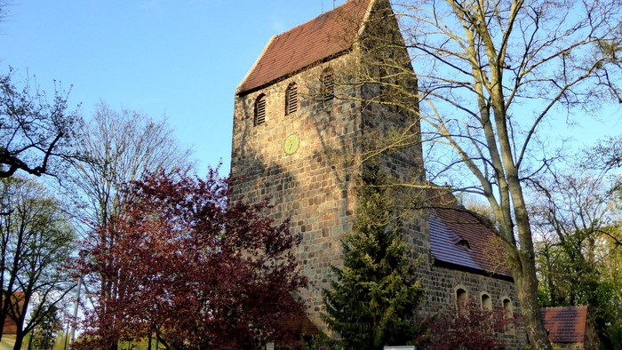 Alte Kirche mit hohem Turm und rotem Dach, umgeben von Bäumen und Büschen