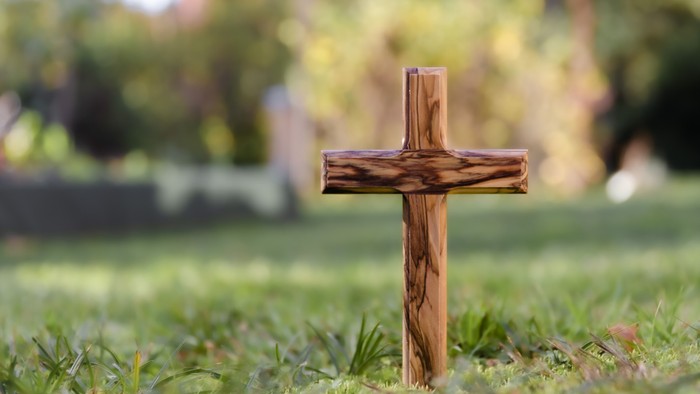 Ein hölzernes Kreuz ragt deutlich auf einem grasbewachsenen Friedhof mit unscharfen Hintergrundbäumen hervor.