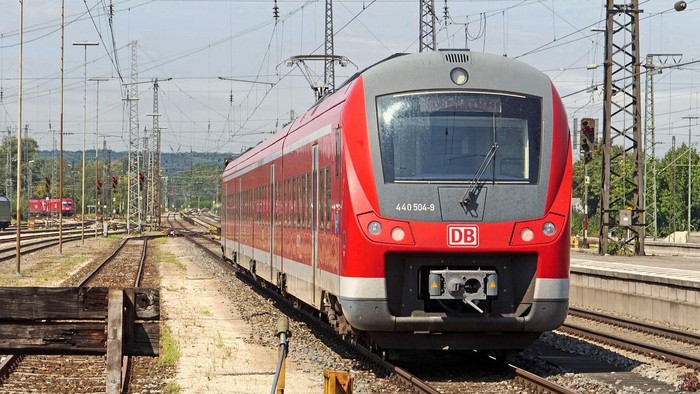 Ein roter und silberner DB-Personenzug fährt auf elektrifizierten Gleisen durch eine ländliche Gegend.