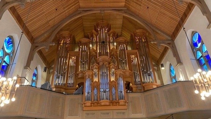 Elegantes Pfeifenorgel in einer Kirche mit kunstvoller Holzarbeit und farbenfrohen Buntglasfenstern dahinter.