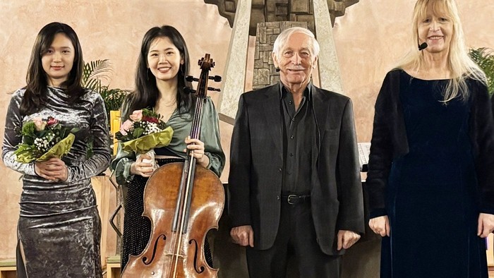 Vier Personen stehen nebeneinander in einem Kirchenraum oder Konzertsaal: zwei Musikerinnen mit Blumensträußen (eine davon mit Cello), ein älterer Mann im Anzug und eine Frau mit Headset-Mikrofon; alle blicken in die Kamera, im Hintergrund ist eine künstl