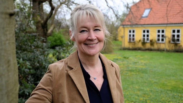 En smilende kvinde i en brun blazer står udendørs nær en charmerende gul bygning med grønne græsplæner.