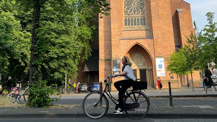 Ein Radfahrer fährt an einer hohen Backsteinkirche mit einem spitzen Turm und bunten Glasfenstern vorbei.