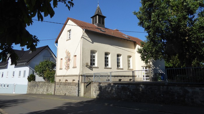 Idyllische weiße Kirche mit kleinem Turm und rotem Dach, eingebettet zwischen belaubten Bäumen.