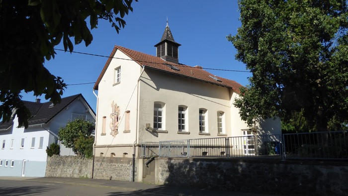 Idyllische weiße Kirche mit kleinem Turm und rotem Dach, eingebettet zwischen belaubten Bäumen.