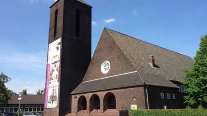 Kirche mit hohem Turm und Kreuzsymbol an der Fassade, Banner hängt vom Turm herab, blauer Himmel im Hintergrund.
