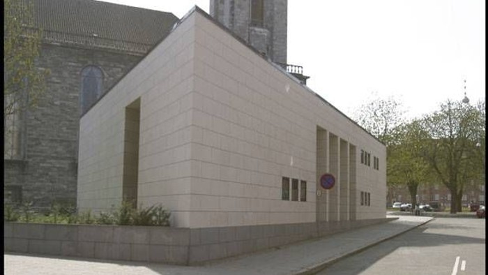 Et moderne bygning med kubisk form og flade vegger, placeret ved en gade med træer og en kirke i baggrunden.