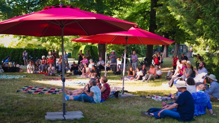 Menschen versammeln sich im Freien unter einem roten Schirm und genießen ein sonniges Picknick in einer grünen Parklandschaft.