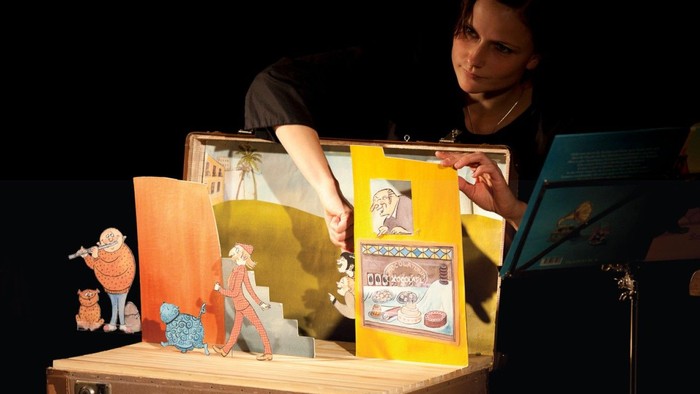 Ein*e Künstler*in nutzt ein Kofferpuppentheater, um zu unterhalten – mit bunten Szenen und Figuren darin.