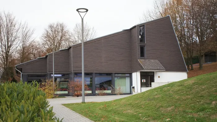 Modernes Gebäude mit schrägem Dach und großen Fenstern, das auf einem grasbewachsenen, gestalteten Hang liegt.