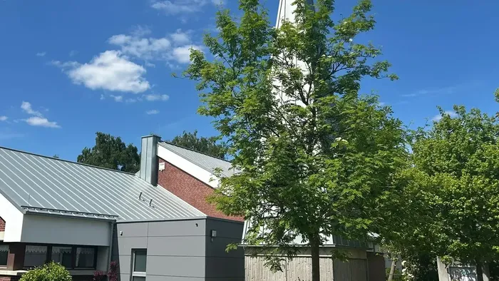 Ein Kirchturm erhebt sich über üppig grüne Bäume unter einem klaren blauen Himmel mit angrenzenden Gebäuden.