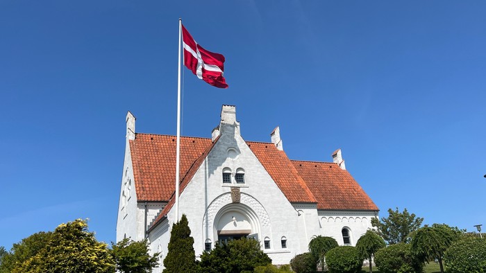 Et malerisk hvidt hus med rødt teglstenstag, hvor et dansk flag vajer under en klar blå himmel.