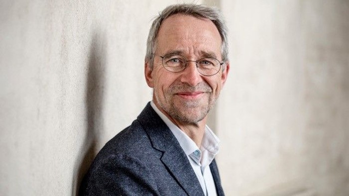 En smilende mand i jakkesæt og briller, der læner sig op ad en lys væg.