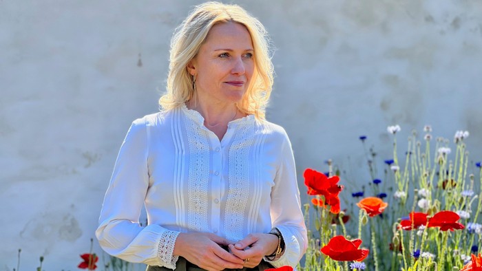 En kvinde i en lys bluse står smilende mellem livlige røde og blå blomster ved en mur.