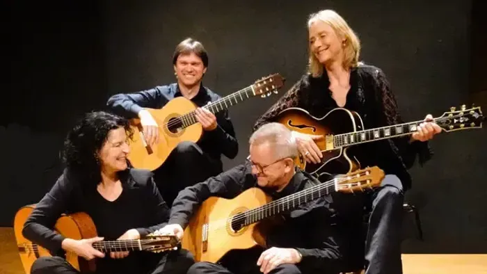 Ein Quartett von Musiker:innen, die auf der Bühne verschiedene Gitarren spielen, sitzend und lächelnd.