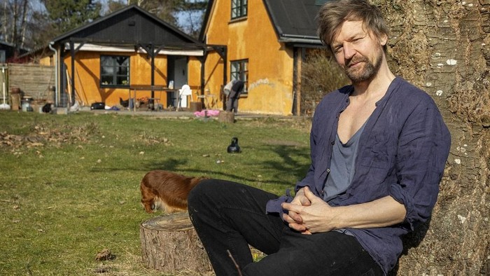 En mand sidder på en stub med en hund ved siden af et idyllisk gult hus i en fredelig baghave.