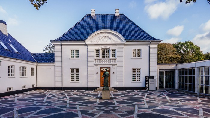 Elegant hvid bygning med blåt tag og et mønstret gårdanlæg med en statue og geometrisk belægning.