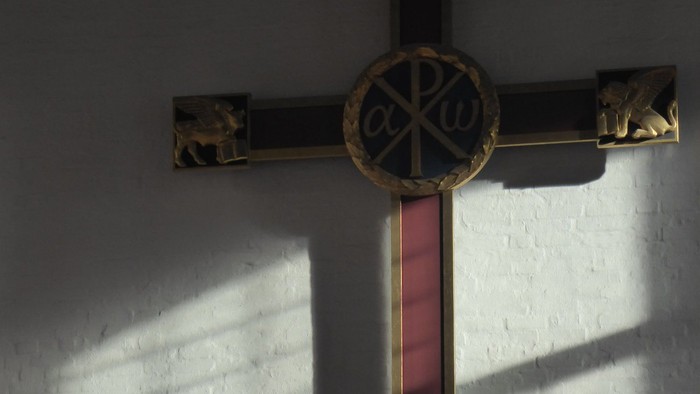 Guldfarvet kors med rød stang og symbol i midten.