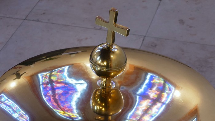 Goldenes Kruzifix auf einem spiegelnden Taufbecken mit farbigen Glasmalerei-Reflexionen.