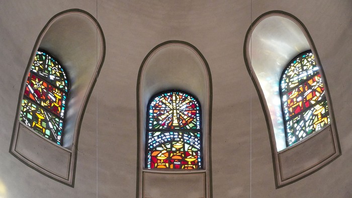 Drei bogenförmige Buntglasfenster mit farbenfrohen religiösen Motiven in einer beigen Wandnische.