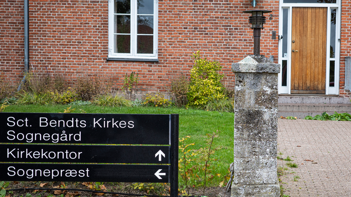 Skilt, der viser vej til Sct. Bendts Kirke, dens have, kontor og præstegård med indgang til murstensbygning.