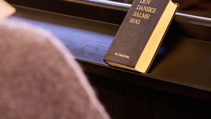 En salmebog med titlen *Den Danske Salmebog* placeret på en kirkebænk i en kirke.
