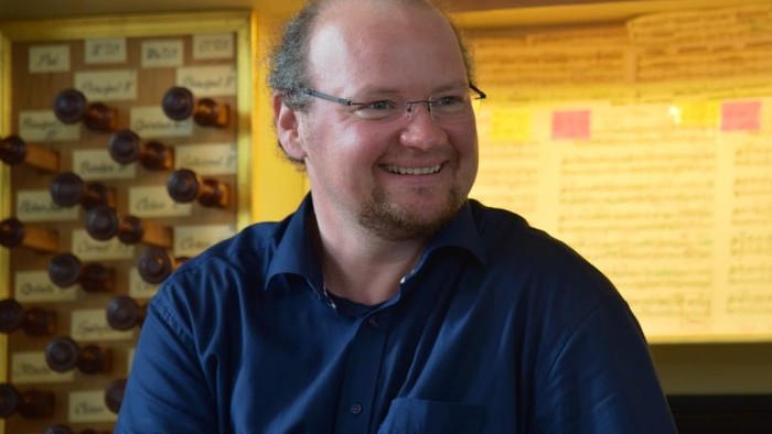 Mann mit Brille sitzt vor einem Weinregal und einem gelben Brett mit Notizen.