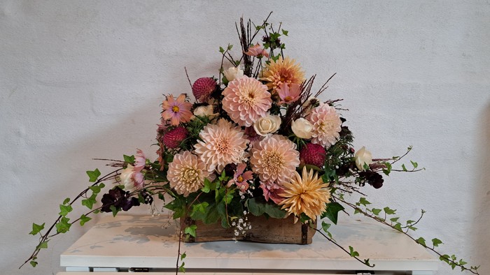 Elegant blomsterarrangement i en rustik trææske med bløde pastelfarvede blomster og grønt på en glat hvid overflade.