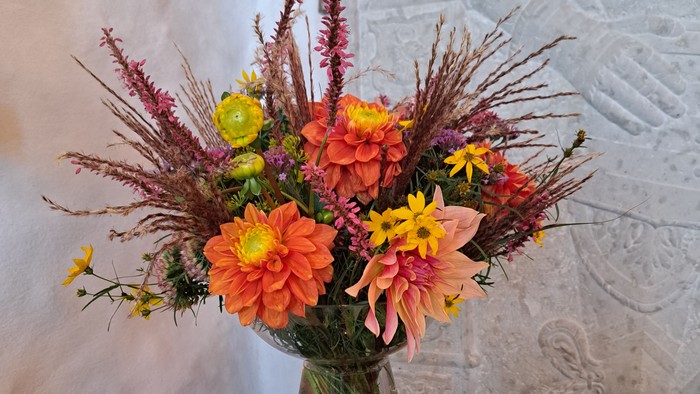 Levende buket i en gennemsigtig vase med orange, gule og lilla blomster foran en struktureret væg.