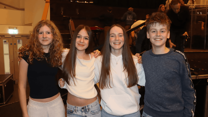 Four smiling teens pose together in a brightly-lit stage or performance area.
