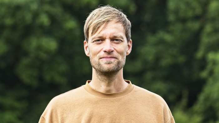 En person med kort, blondt hår iført en beige sweater på en baggrund af grønt løv.