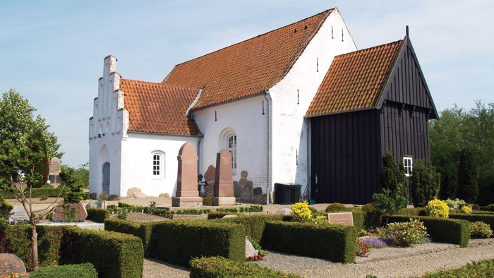 Et hvidt kirkebygning med røde tag og en træbygning til højre for, omgivet af en have med buske og blomster.