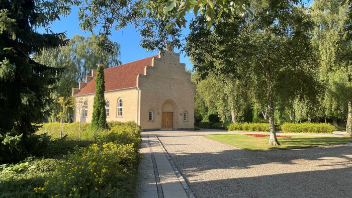 En lille kirke med rødt tag og trædør, omgivet af grønne træer og buske